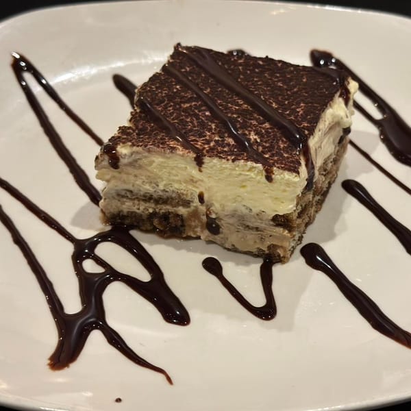 A slice of tiramisu with chocolate drizzle and cocoa powder on a white plate.