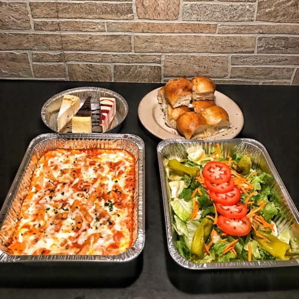 Amore Pasta & Pizza catering spread with baked ziti, garden salad, bread rolls, and cheesecake slices.